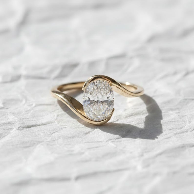 Oval cut diamond bypass engagement ring with asymmetrical open wrap gold band and modern solitaire setting.