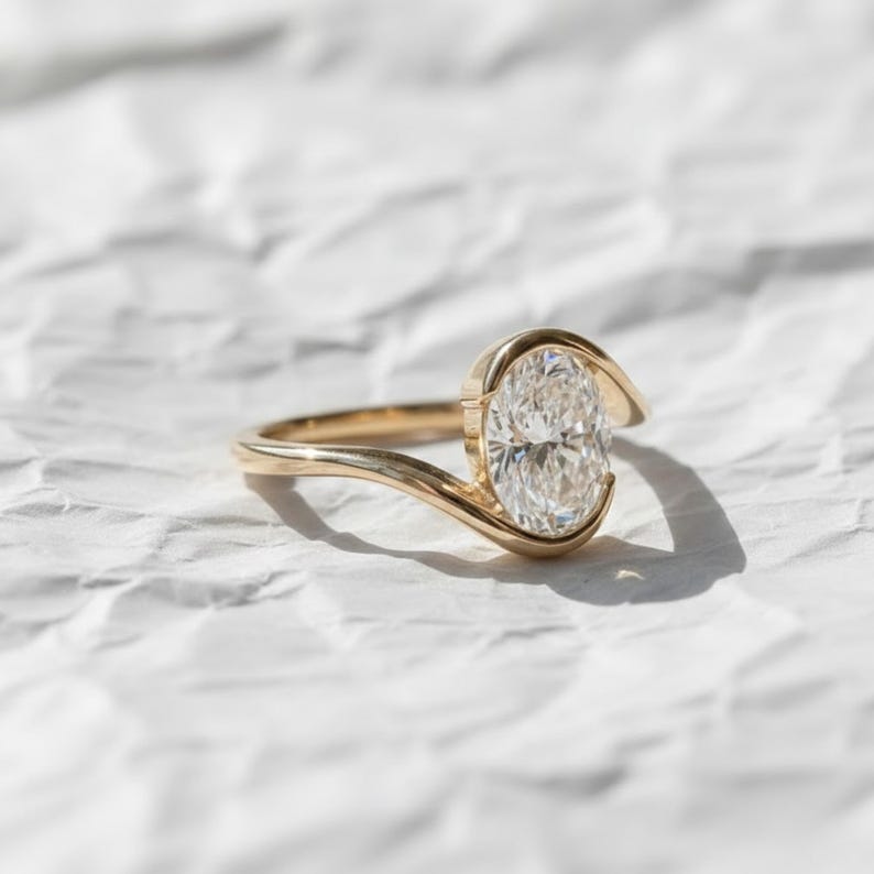 Oval cut diamond bypass engagement ring with asymmetrical open wrap gold band and modern solitaire setting.