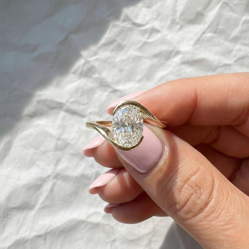 Oval cut diamond bypass engagement ring with asymmetrical open wrap gold band and modern solitaire setting.